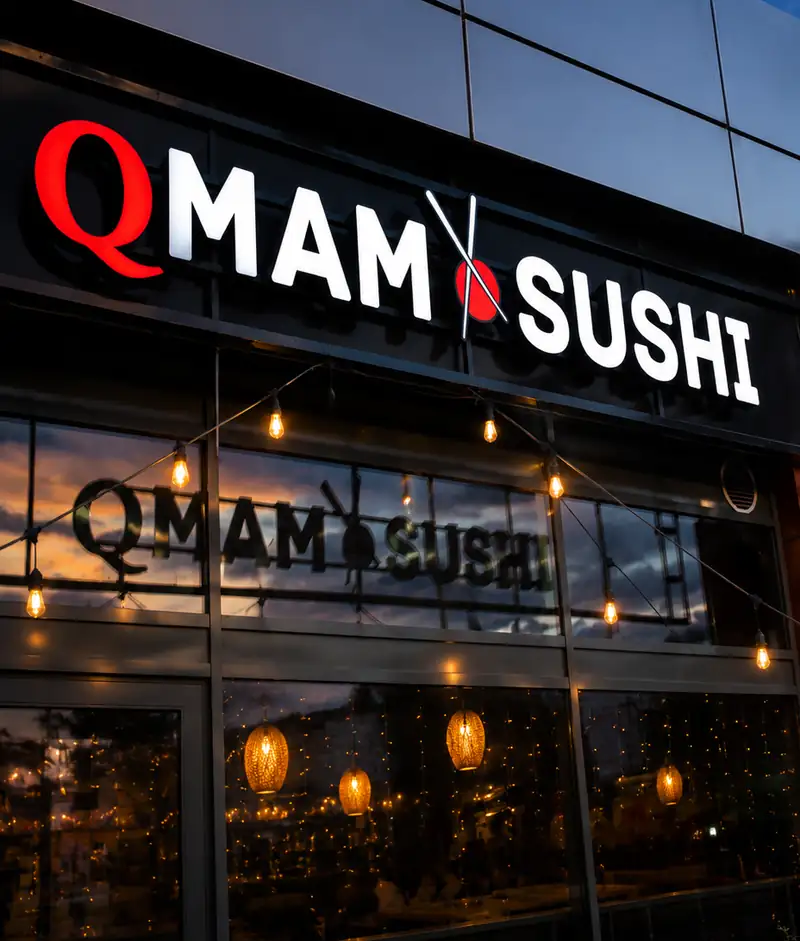 Szyld QMAM SUSHI nad przeszklonym wejściem wieczorem